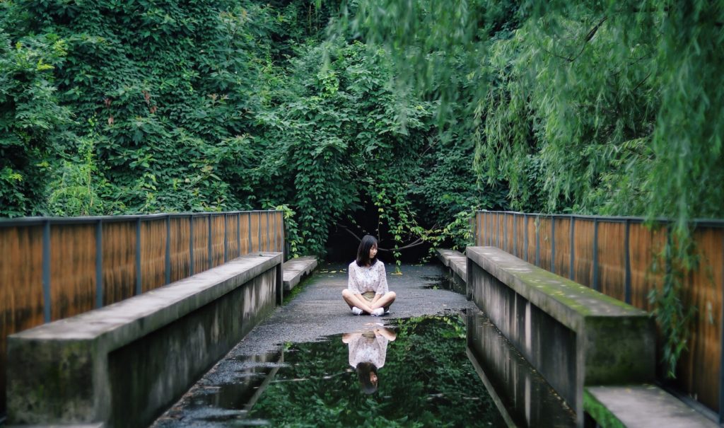 A woman sits on a path surrounded by green foliage. A puddle in front of her reflects her image. Internalized sexism.