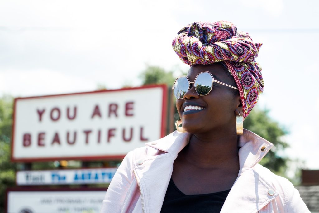 A black woman with sunglasses and a colorful headscarf smiles, posing in front of a sign that says "You are beautiful." Be your own best friend.