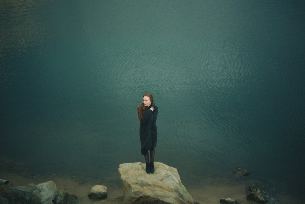 A woman stands small and alone on a rock in front of a body of water stretching out behind her, clothing a blanket to her body. grief in a pandemic