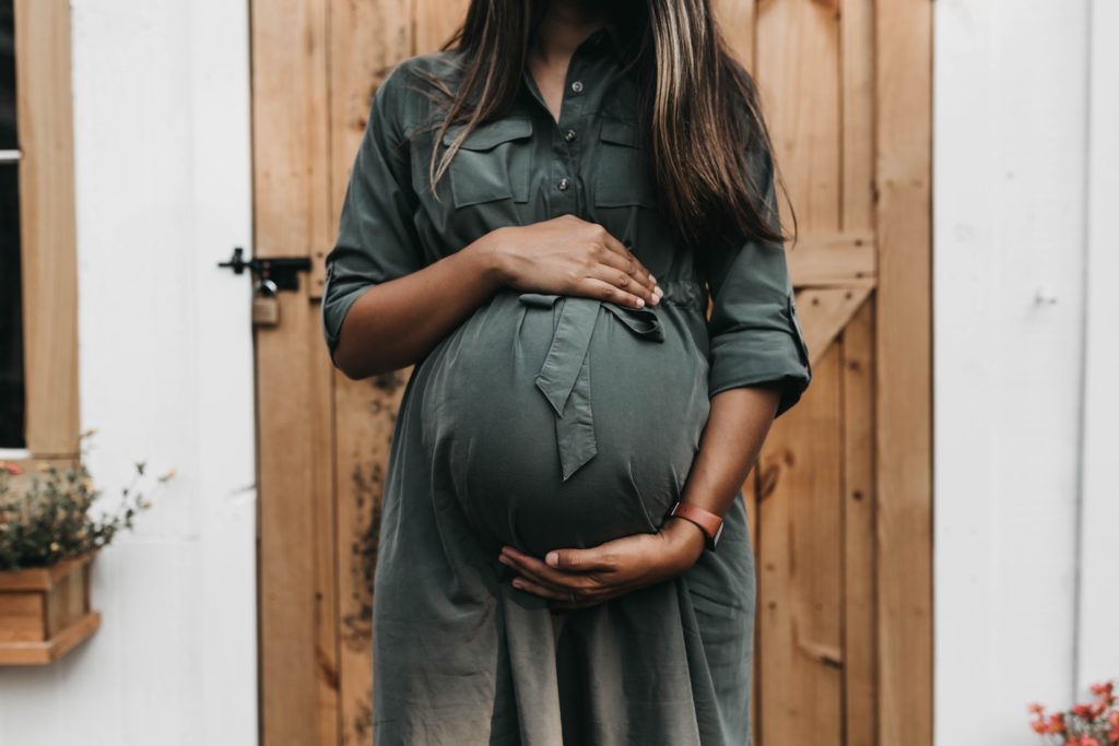 A pregnant woman in an olive green dress stands in front of a wooden door, hands cradling her belly. financial advice and money shame