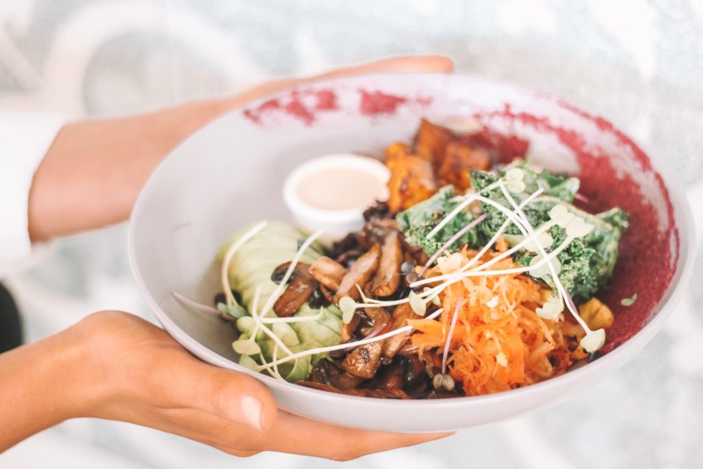 cost of a plant-based diet. hands hold a bowl with slice avocado, mushrooms, shredded carrots, and kale.