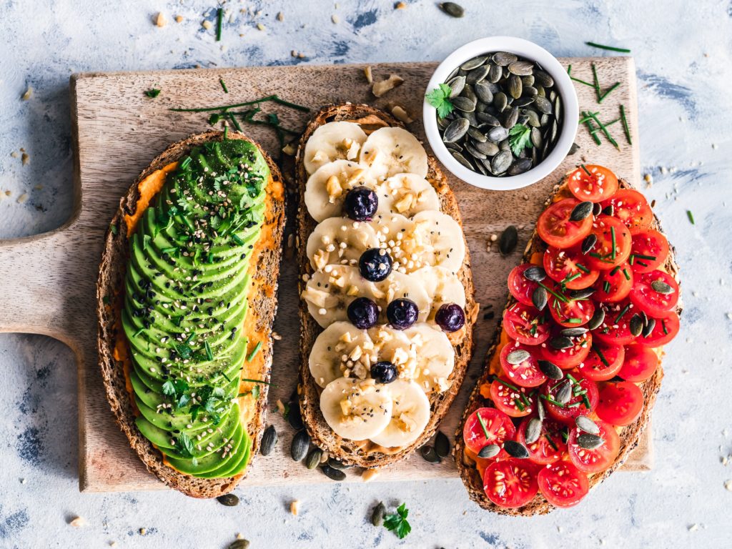 tips to save money on a vegan diet. three toasts sit on a wooden cutting board: an avocado toast, one topped with bananas and blueberries, and another topped with halved grape tomatoes and pumpkin seeds.